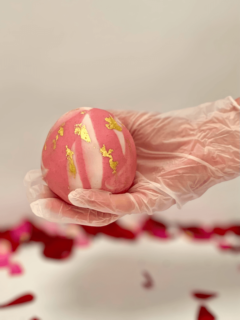 A gloved hand holding a single marbled Chocolate Fudge Cake ball in pink and white decorated with edible gold leaf. 