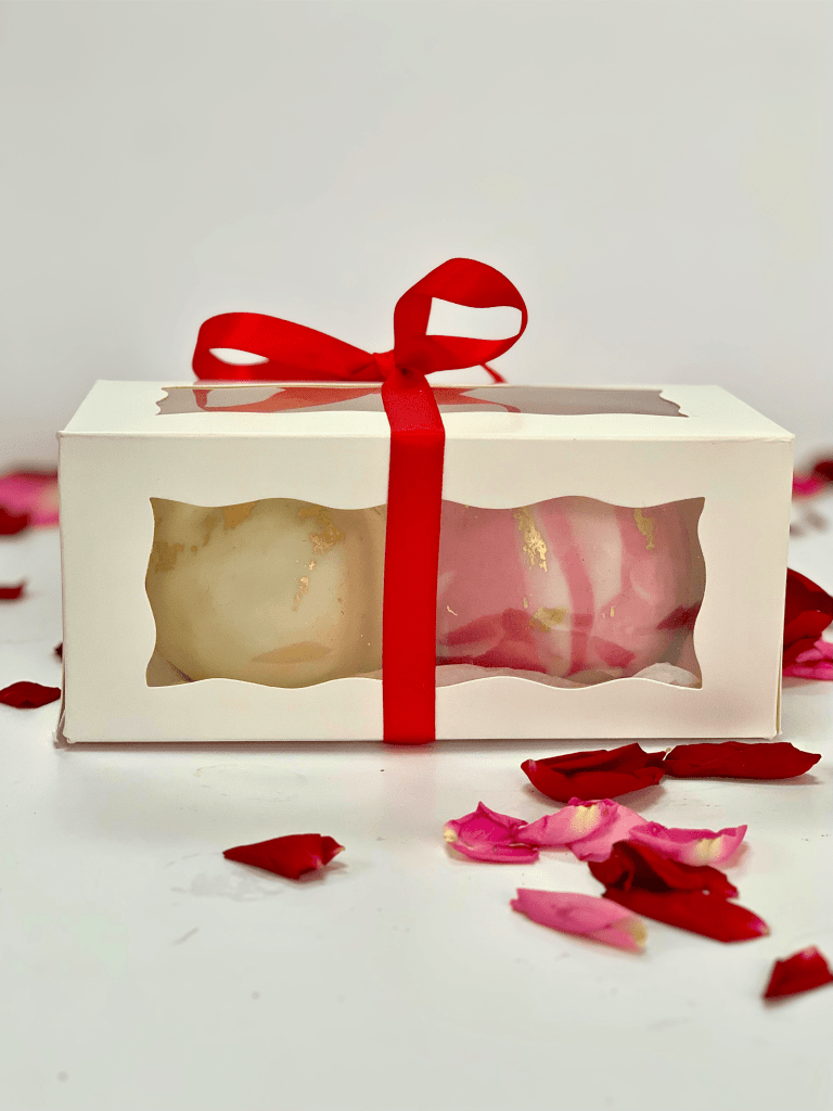 A photo of a Valentine's white gift box containing two chocolate cake fudge balls in marbled fondant and edible gold leaf. The yellow marbled ball is Passion fruit and Lemon and the marbled pink ball is Raspberry & Marc de Champagne flavour. The box is decorated with a red ribbon and bow. 