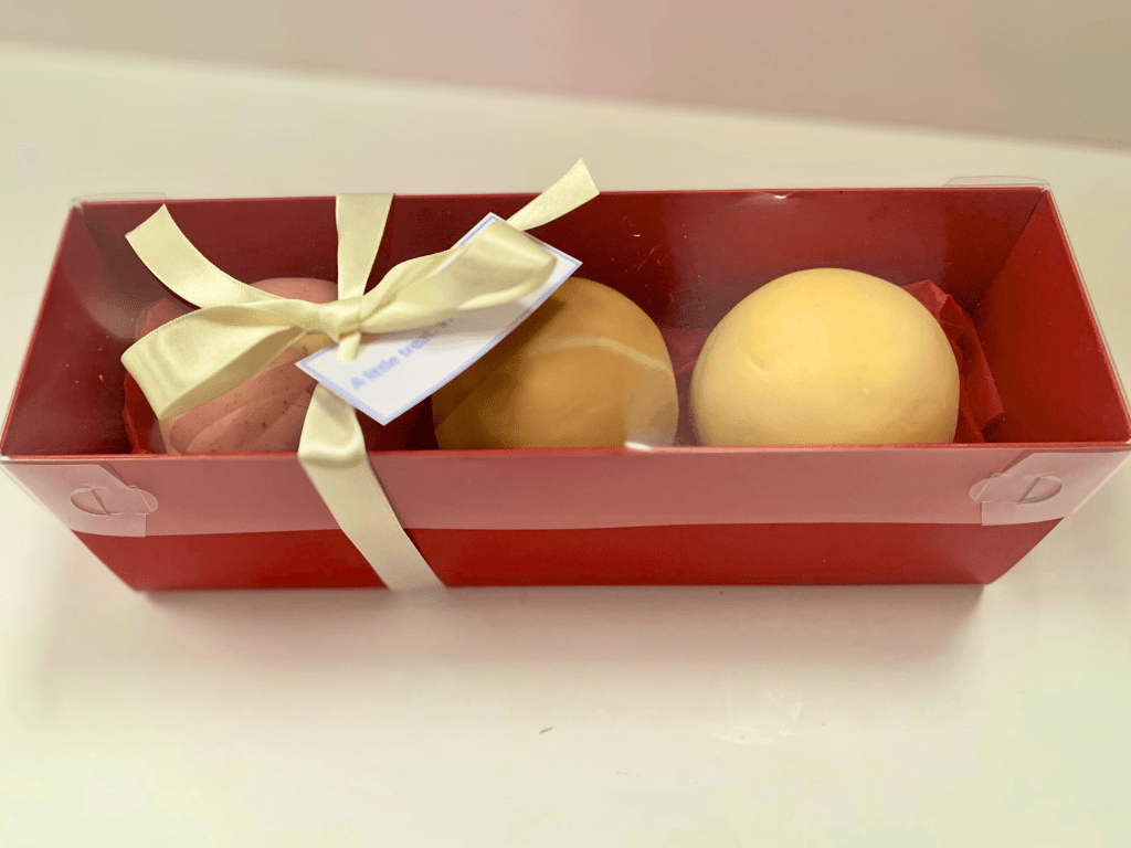 A red gift box of 3 Chocolate Fudge Cake Balls by Cocoa & Whey Cakes. The balls are marbled in pink, brown and yellow fondants. The box is tied with an ivory ribbon with a bow and gift message. 