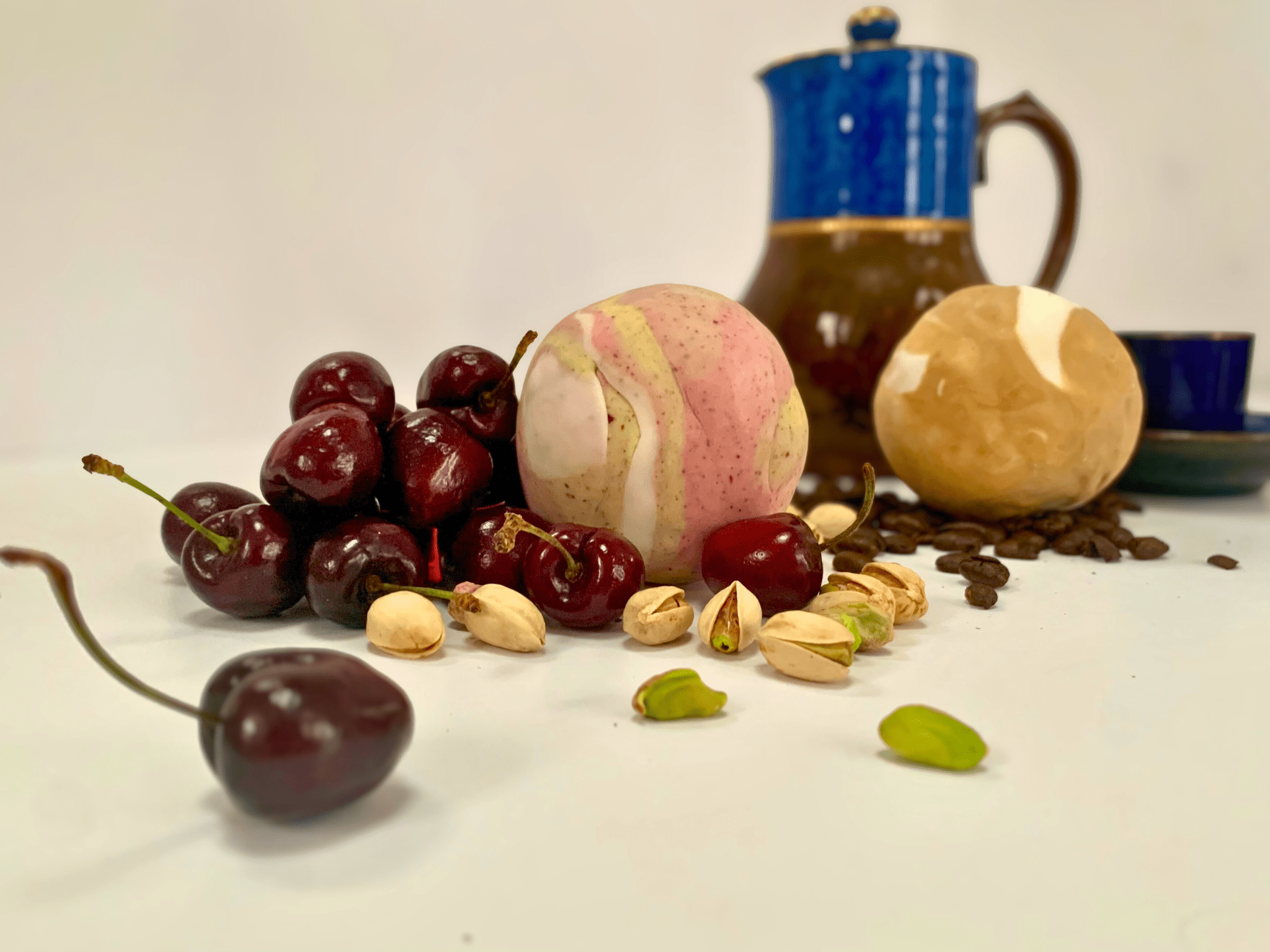 A photo of a table surface with two chocolate cake fudge balls in pink and green as well as browns. There are cherries, pistachio nuts and coffee beans around the cake balls and a china coffee pot in blue, brown and gold behind them. 