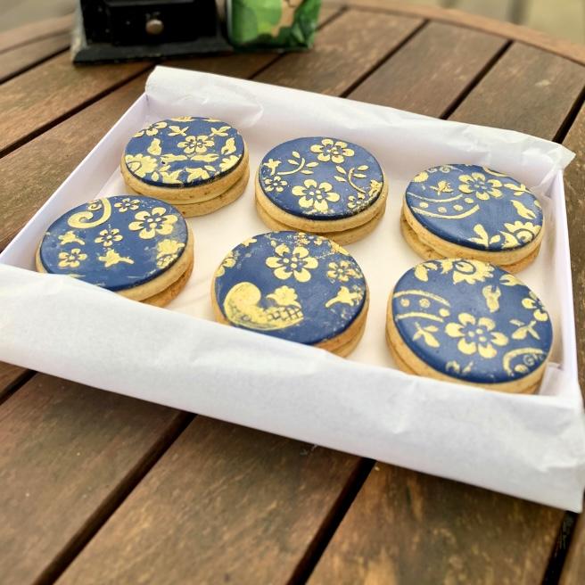A photo of an open white gift box containing 6 double cookies filled with chocolate ganache in Arabica Coffee flavour. The cookies are covered with blue fondant and stencilled with edible gold in a floral design. 