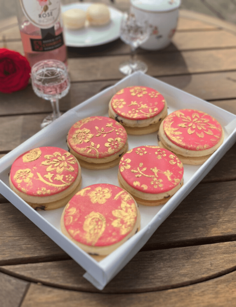 A photo of an open white gift box containing pink double ganache filled Cherry & Rose cookies by Cocoa & Whey Cakes, decorated with edible gold stencilling in a floral design. 